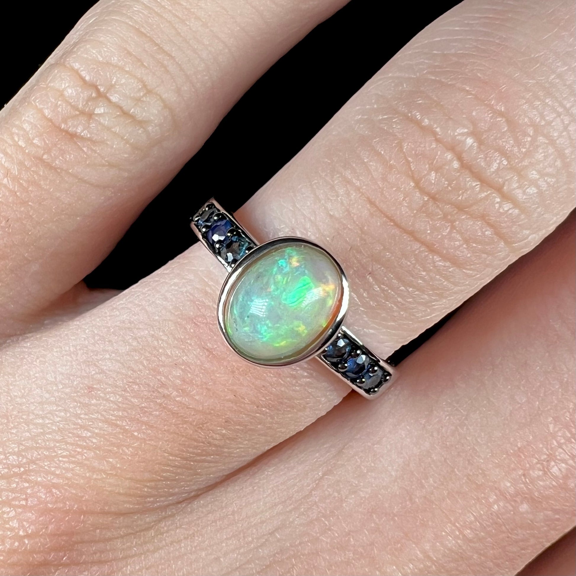 Macro top-down shot of a dainty Ethiopian opal ring with dark blue topaz accents worn on a hand.