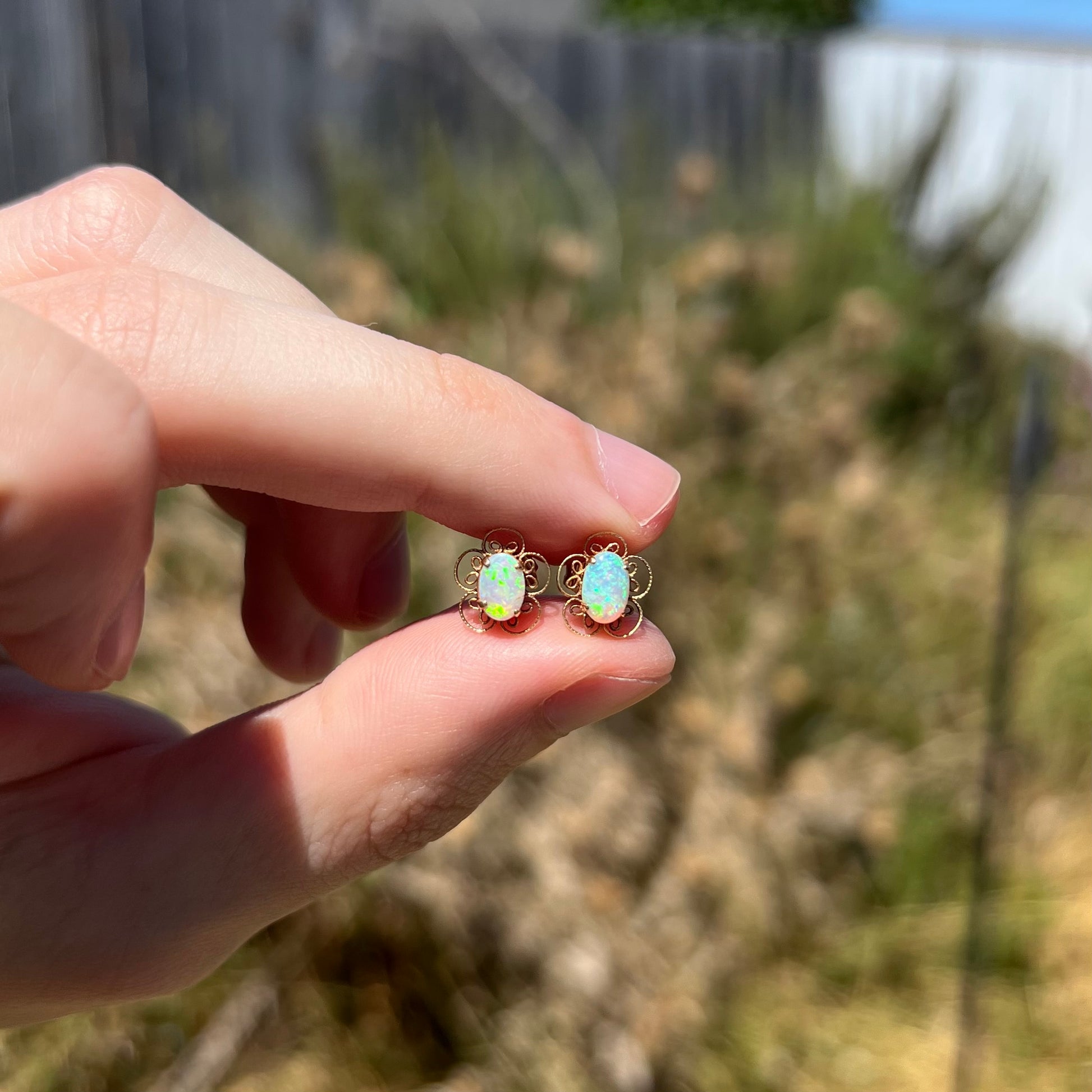 Dainty 14k gold opal flower stud earrings with green flashes of color, held between fingers in sunlight