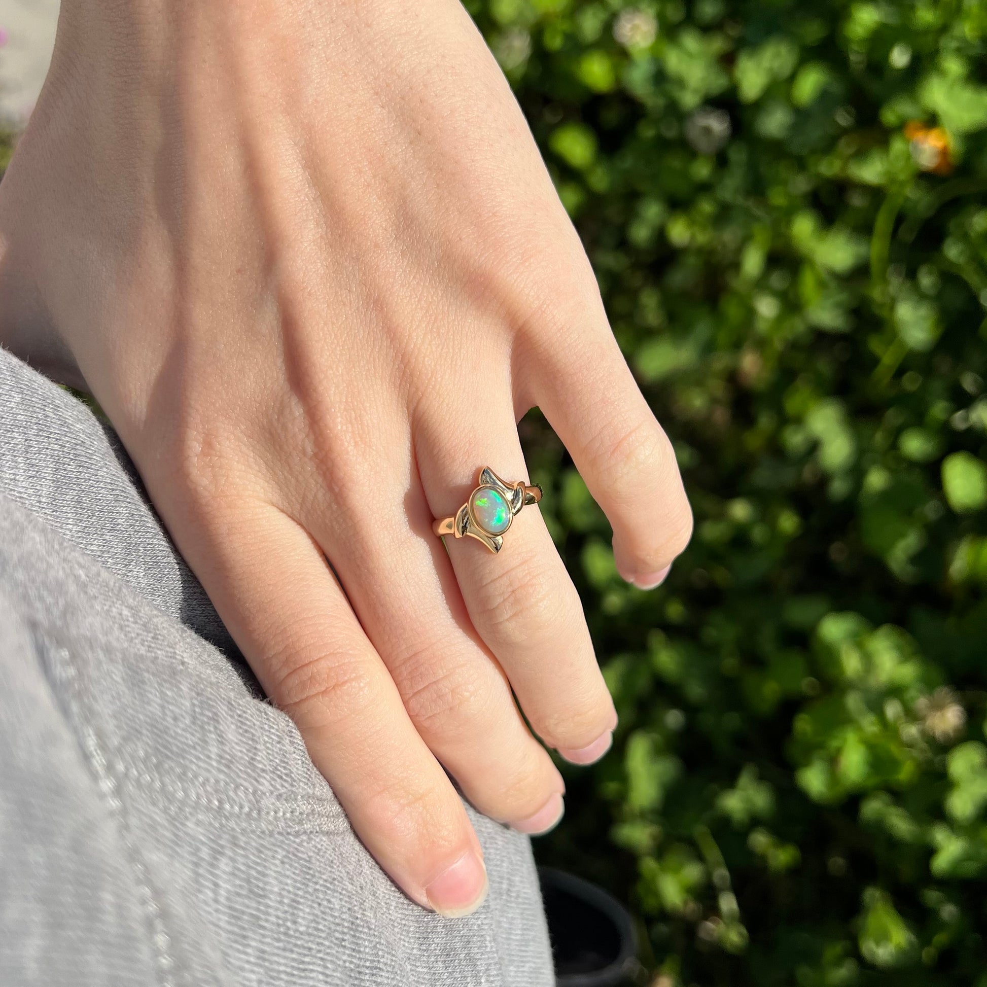 A dainty gold ring set with a green crystal opal, worn on a hand in the sunlight.