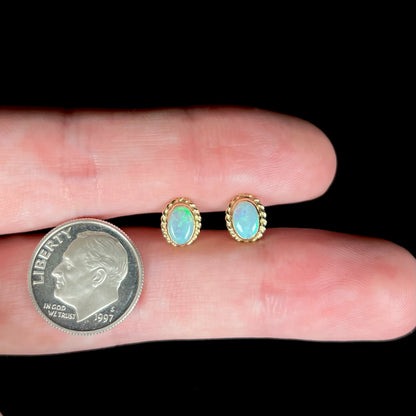 Dainty gold opal stud earrings held between two fingers next to a US dime coin under studio light.