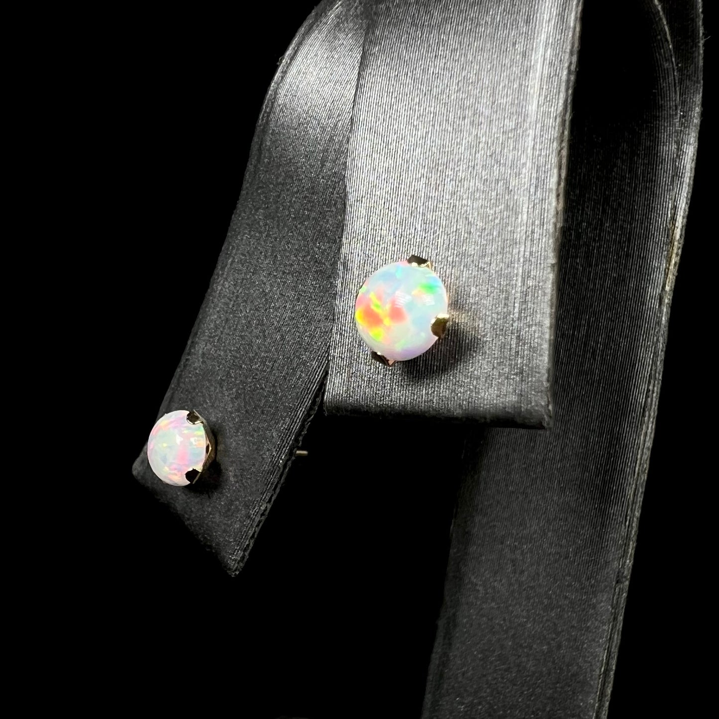 Angled close-up of dainty gold synthetic opal stud earrings in 14k gold, shown on earring display.  The opal in focus has red flashes of color.