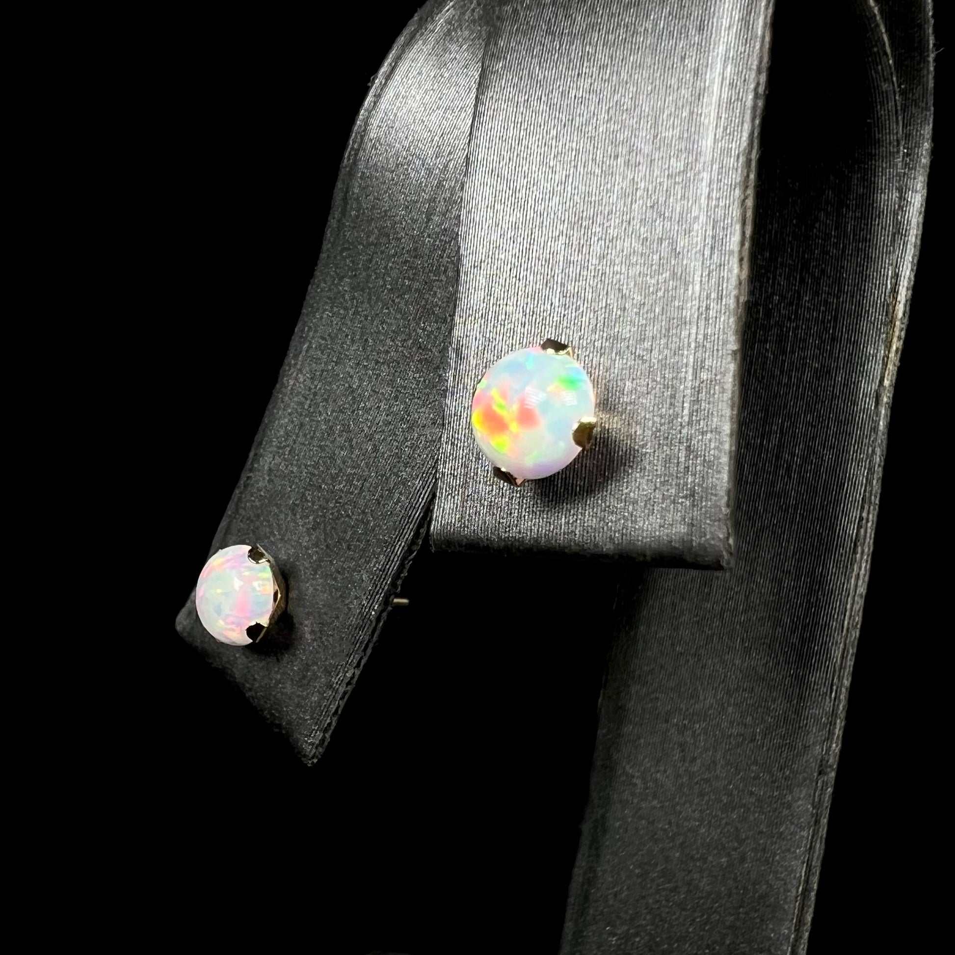 Angled close-up of dainty gold synthetic opal stud earrings in 14k gold, shown on earring display.  The opal in focus has red flashes of color.