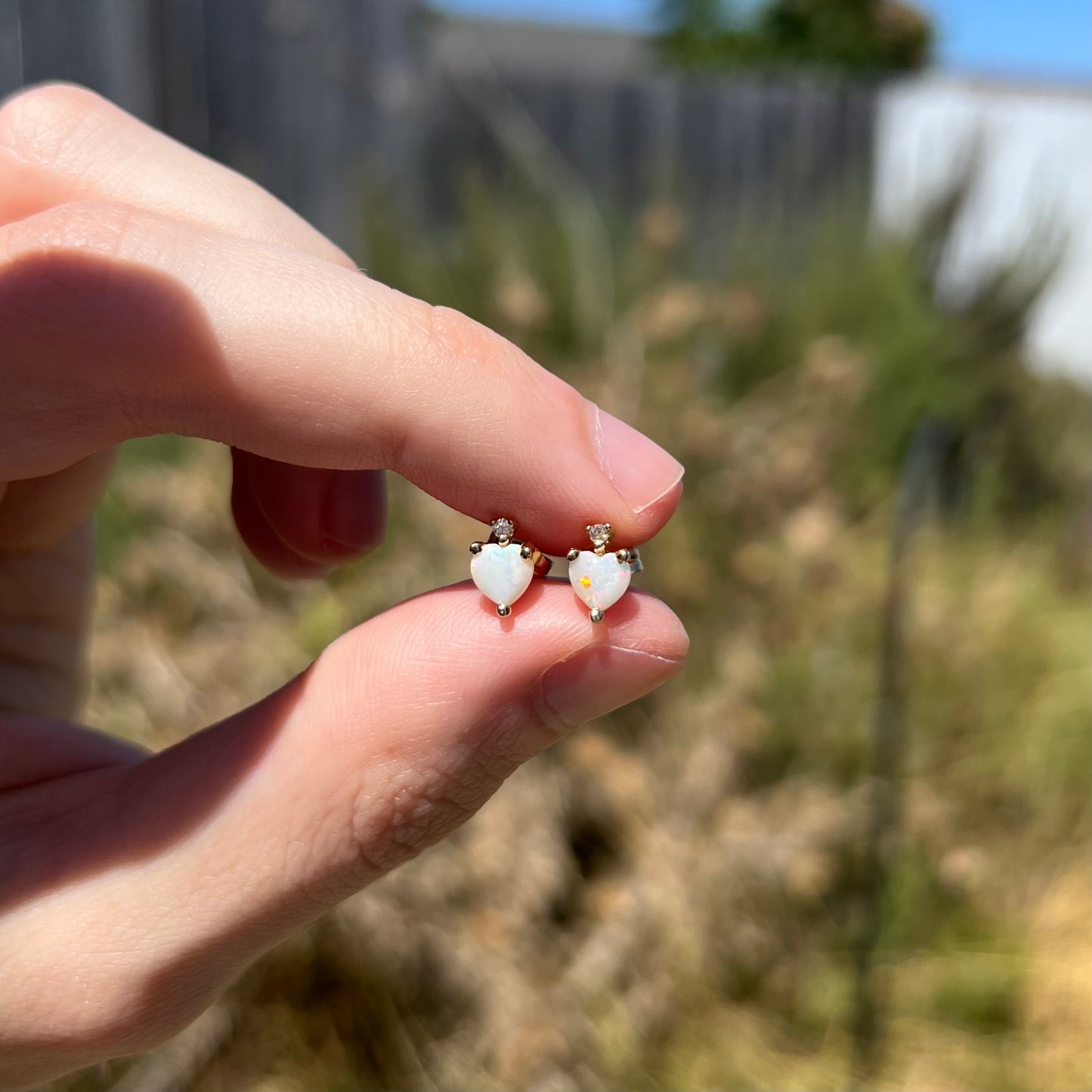 A pair of dainty heart shaped opal stud earrings with diamond accents in hellow gold, held between two fingers in sunlight.