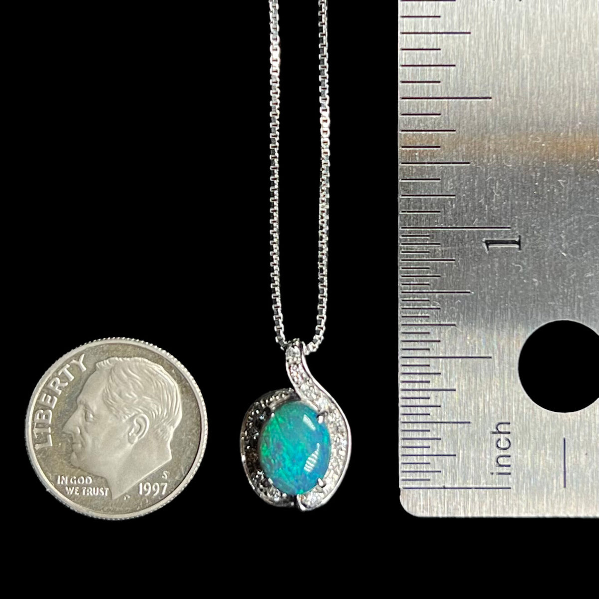 A dainty 1.72ct black opal and diamond pendant that measures two-thirds of an inch in length on chain between US dime coin and ruler for scale.