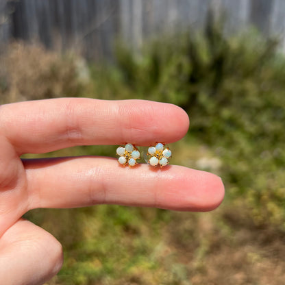 Pair of dainty natural opal flower earrings in gold-tone metal, held between fingers in sunlight