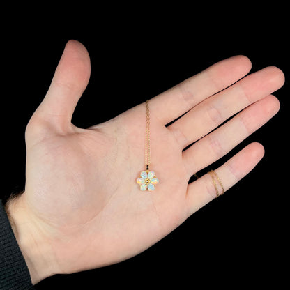Hand holding a dainty women's opal flower pendant fitted to a 14k gold chain; the chain wraps around the pinky finger