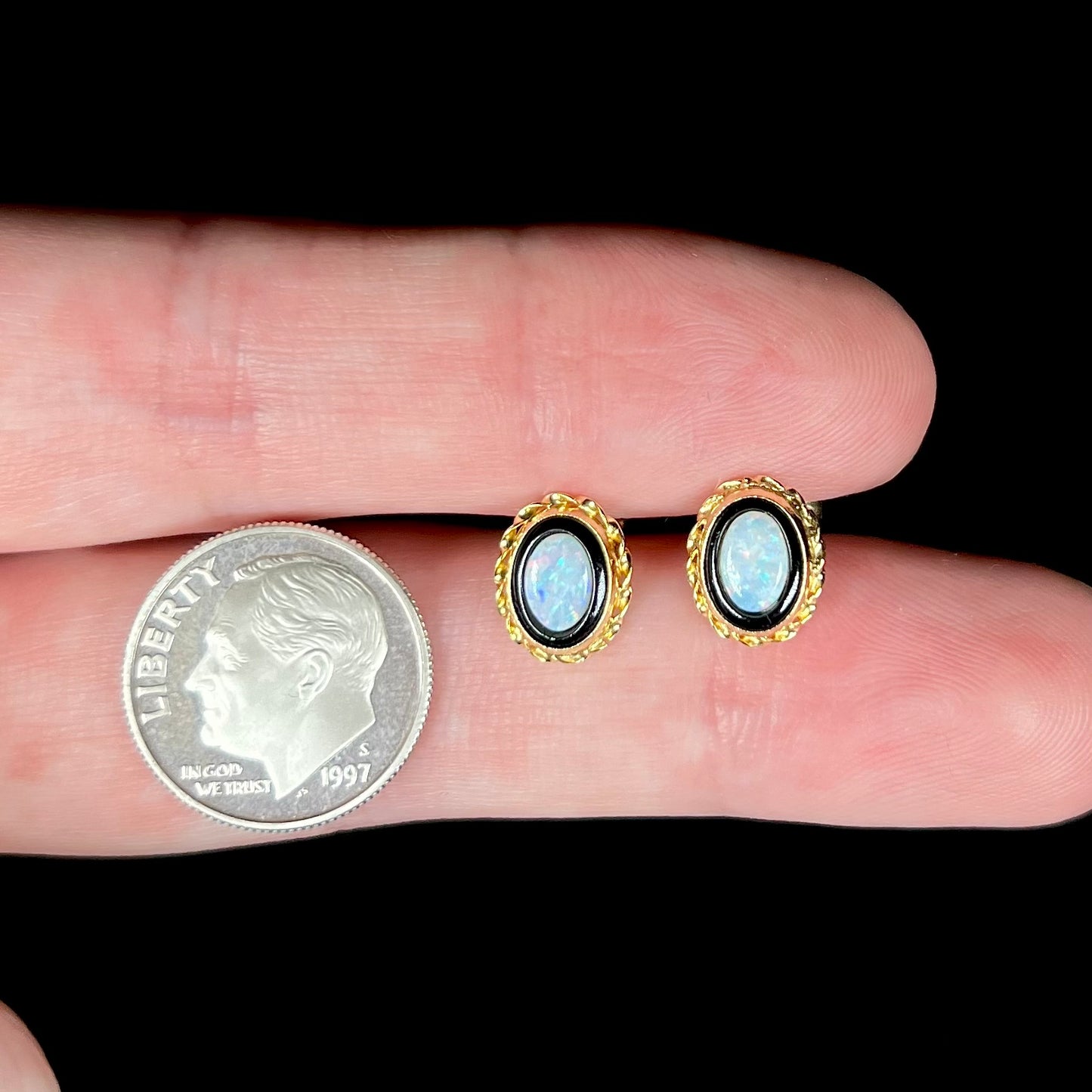 Dainty pair of opal and onyx stud earrings held between two fingers next to a US dime coin.