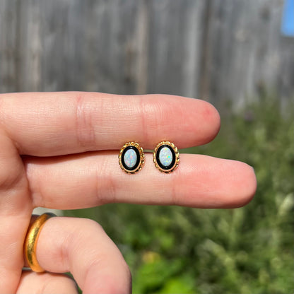 Dainty gold opal and onyx stud earrings held between two fingers in natural sunlight against blurred outdoor background.