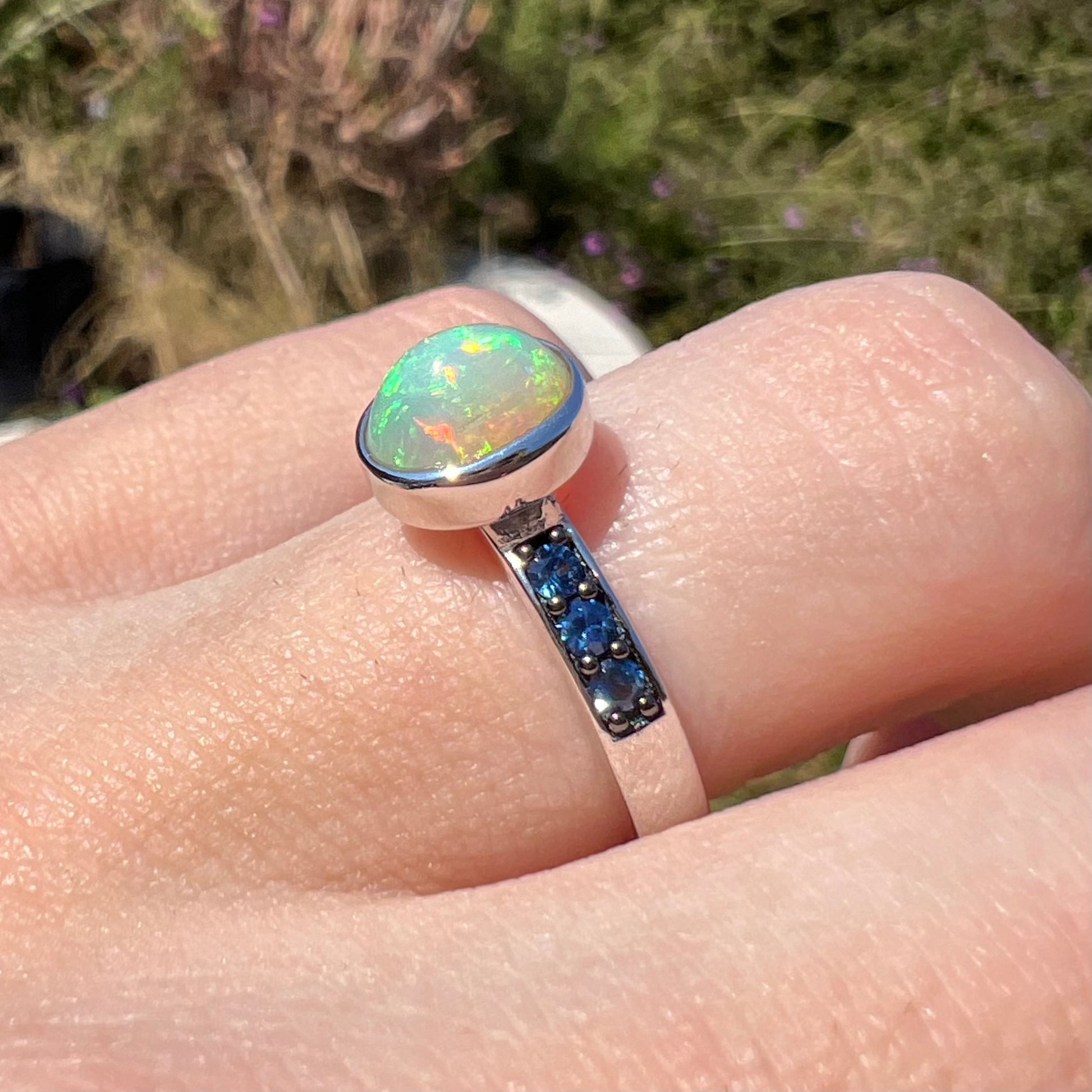 A dainty opal ring worn on a hand in the sunlight, shown at an angle, with the ring's blue accent stones in focus.