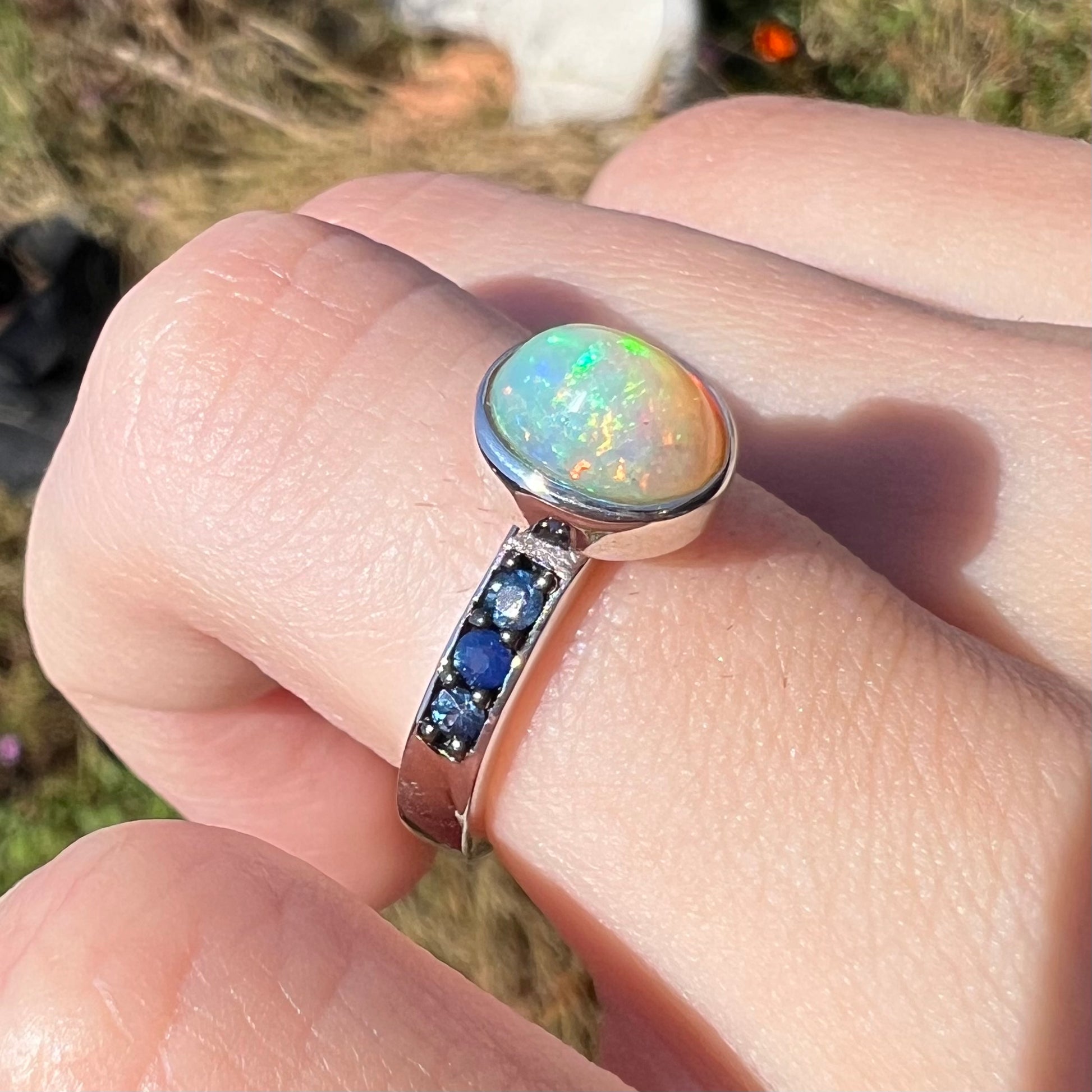 Macro angled view of a dainty opal ring with blue accent stones worn in the sunlight.