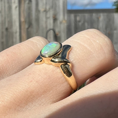 A dainty yellow gold ring set with a Lightning Ridge crystal opal, worn on a hand in sunlight.