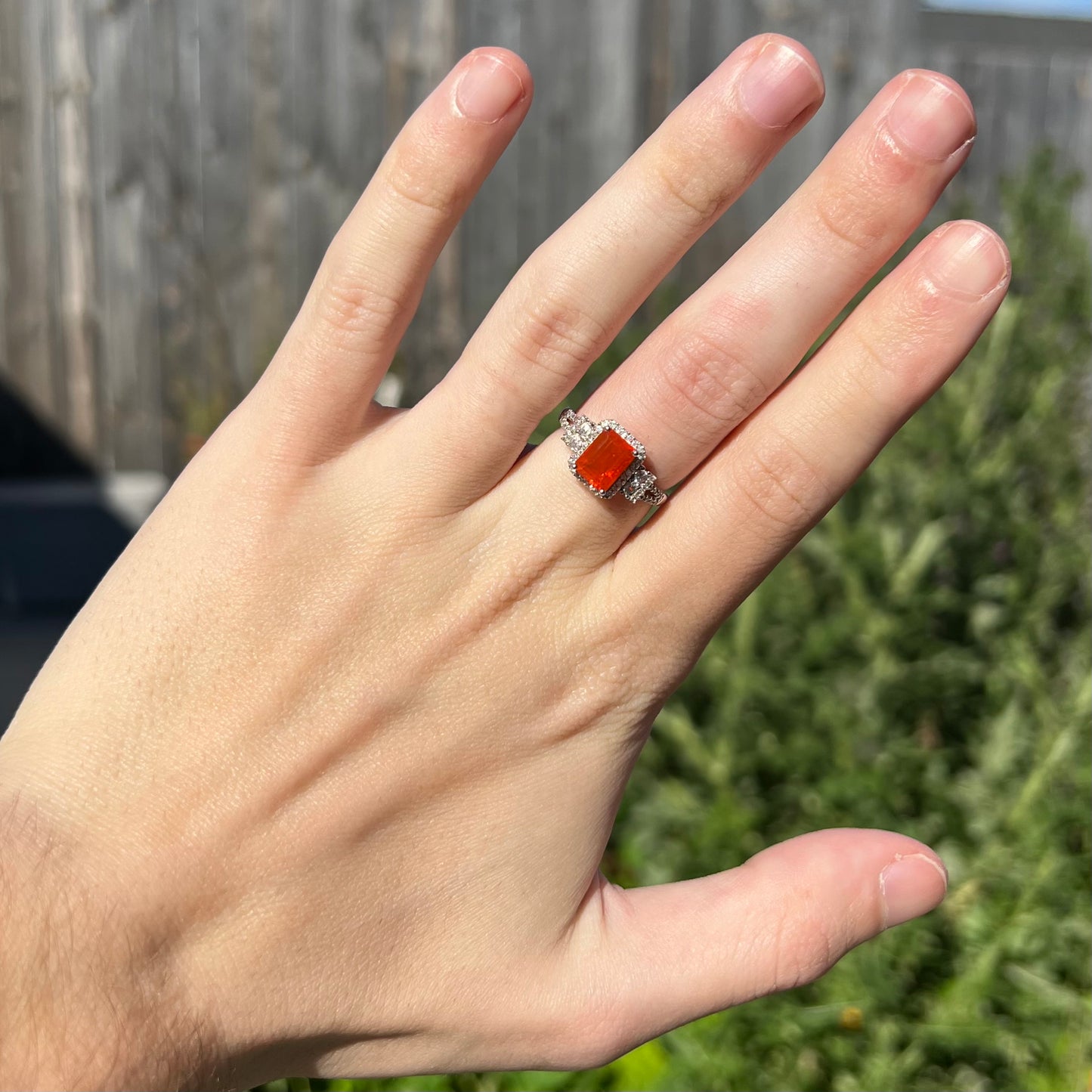 Hand wearing a dainty orange Mexican fire opal ring in natural light agianst a blurred green and brown outdoor backdrop.