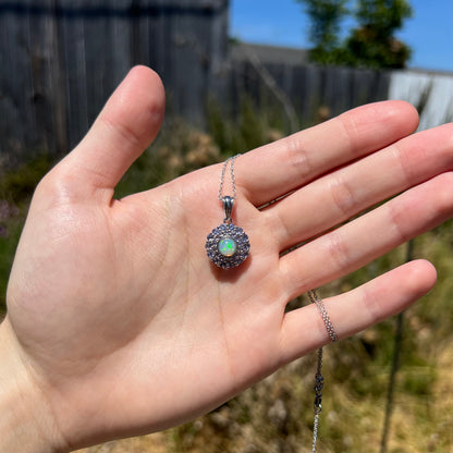 A dainty, round Ethiopian opal and tanzanite halo pendant held under natural sunlight in a hand.