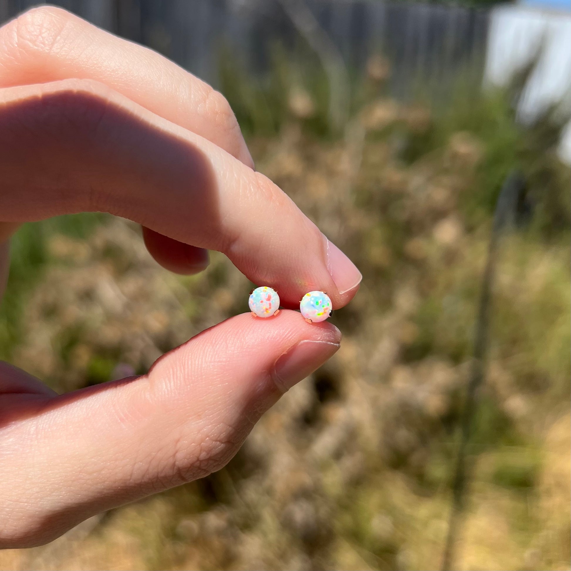 Hand holding a dainty pair of 14k gold round lab-created opal stud earrings between two fingers in sunlight against blurred outdoor backdrop.