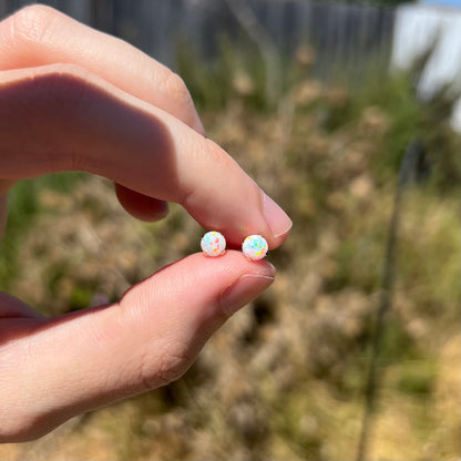 Hand holding a dainty pair of 14k gold round lab-created opal stud earrings between two fingers in sunlight against blurred outdoor backdrop.