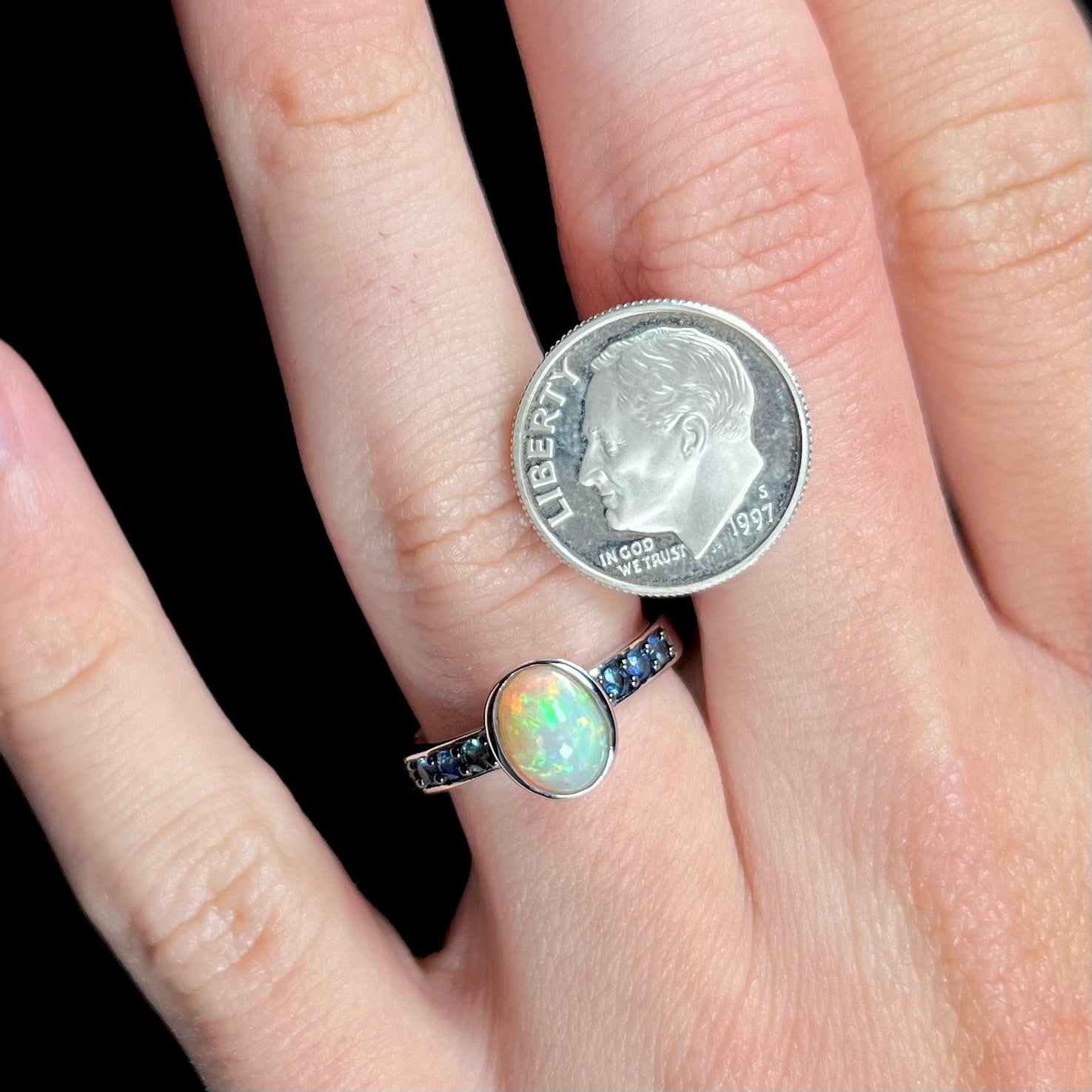 A dainty silver opal ring with blue gemstone accents worn on a hand next to a dime coin for scale.