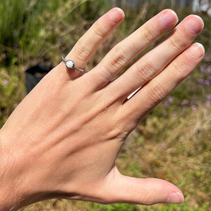 A dainty silver opal ring worn on the pinky finger of a hand in the sun.