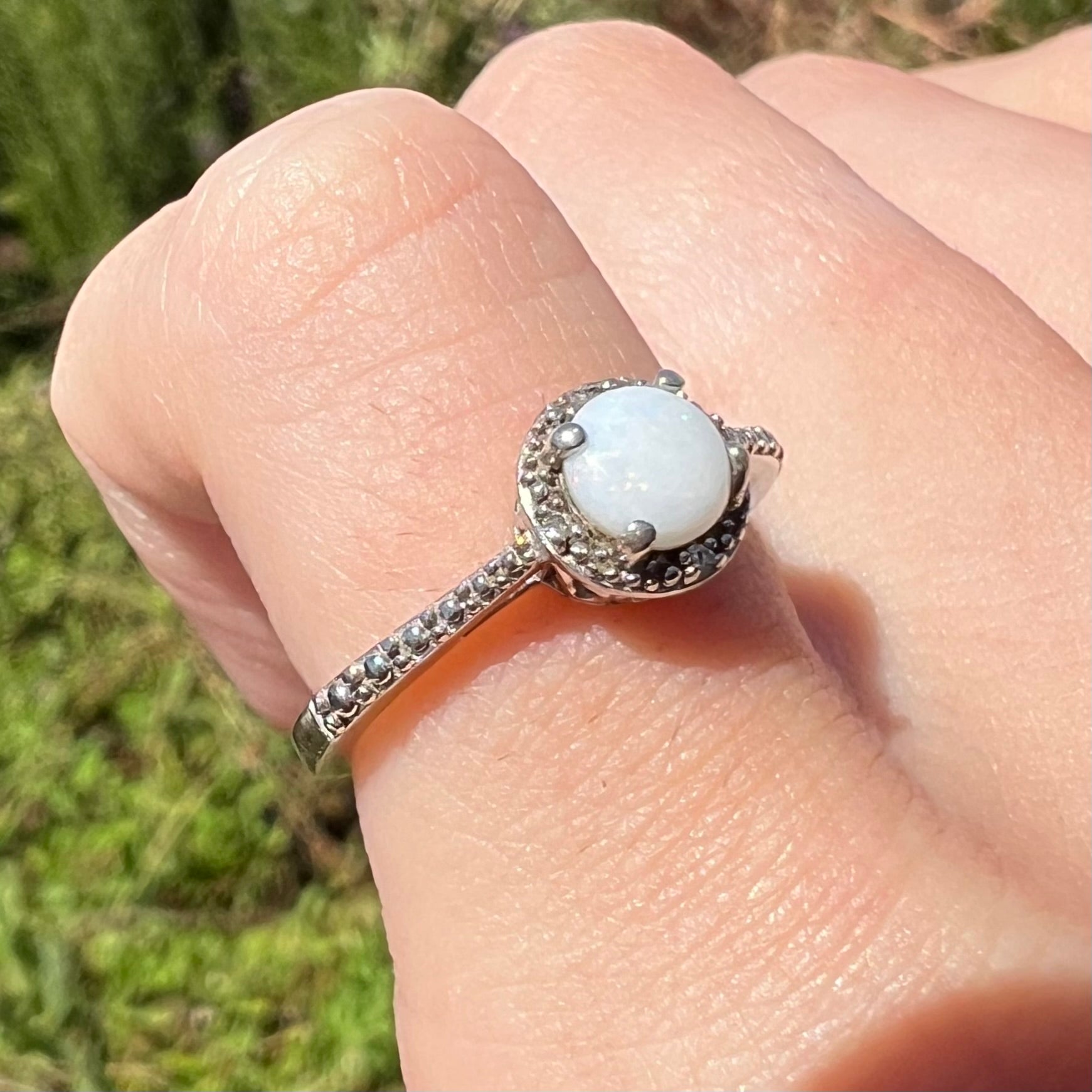 A dainty silver opal ring shown at an anglein natural sunlight, worn on a pinky finger.