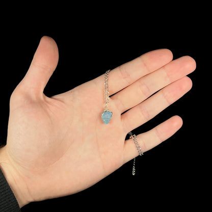 Hand holding a dainty sterling silver wire wrapped dark opal doublet necklace against a black background beneath studio LED light.