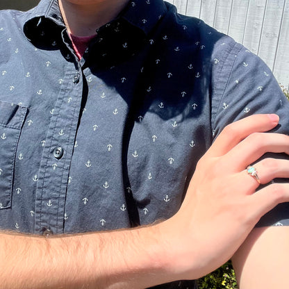 Close-up of person resting hand on opposing shoulder wearing dainty 9k gold Victorian opal ring in sunlight