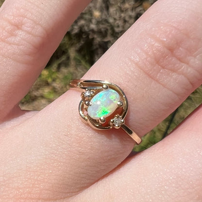 Macro close-up of a vintage gold Australian opal and diamond engagement ring with a dainty design, worn on a finger in natural light.  The opal has green flashes.