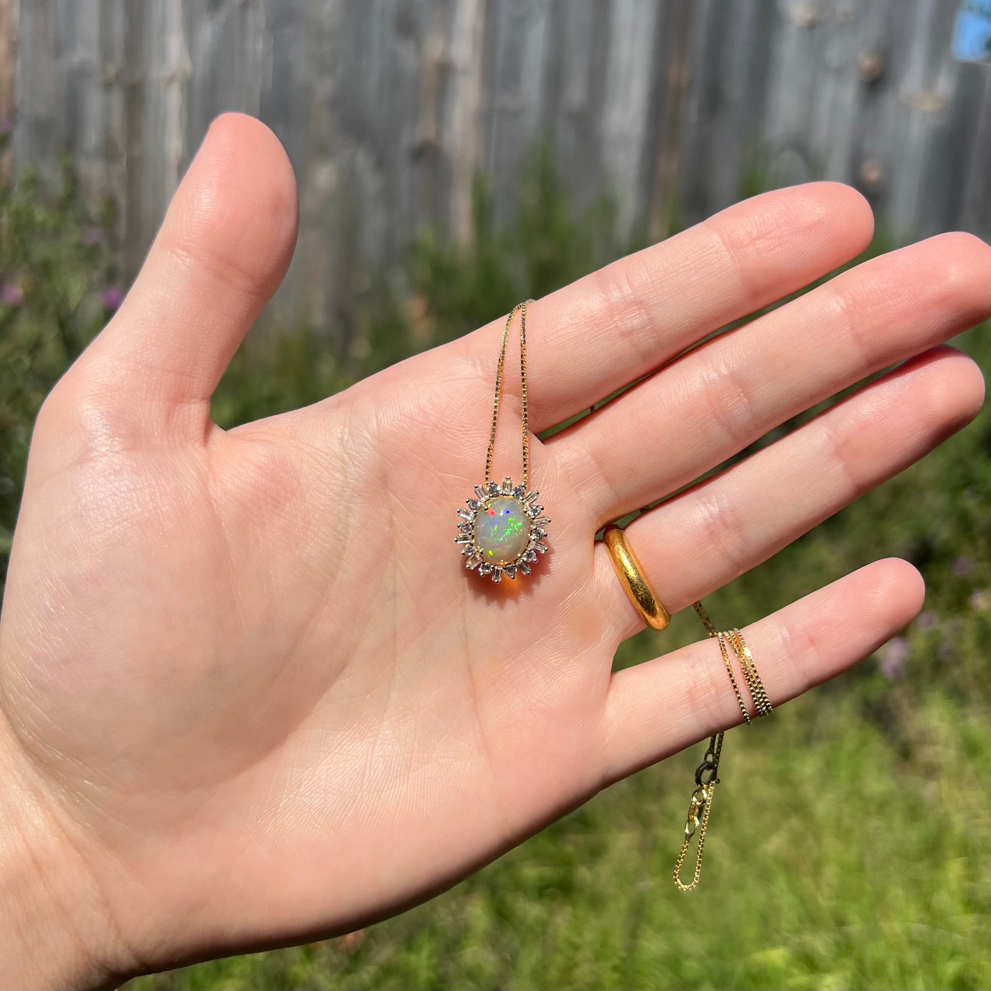 Hand holding a dainty women's 14k yellow gold Ethiopian fire opal and white sapphire halo necklace in sunlight.