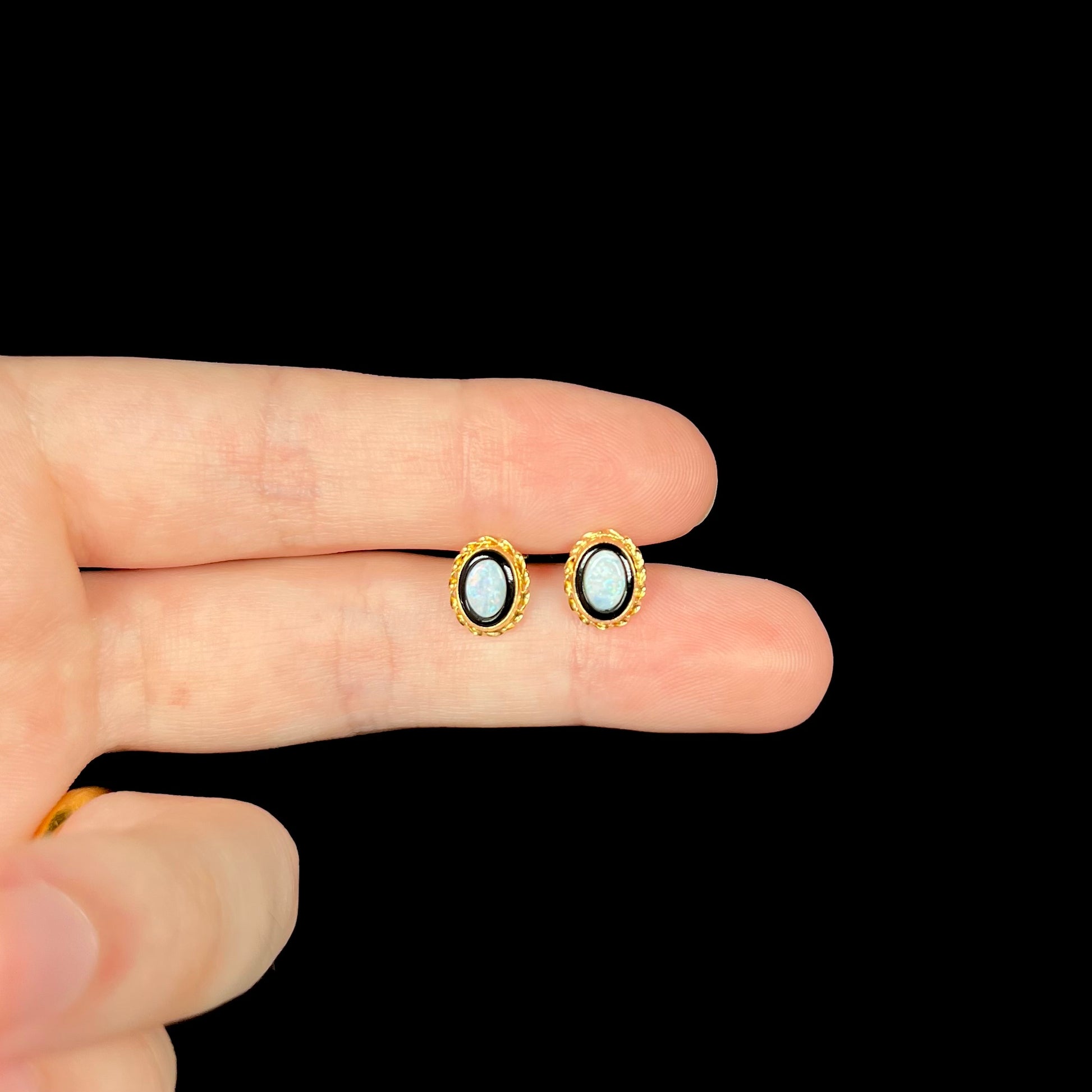 Dainty pair of yellow gold opal in onyx stud earrings held between two fingers against black background.