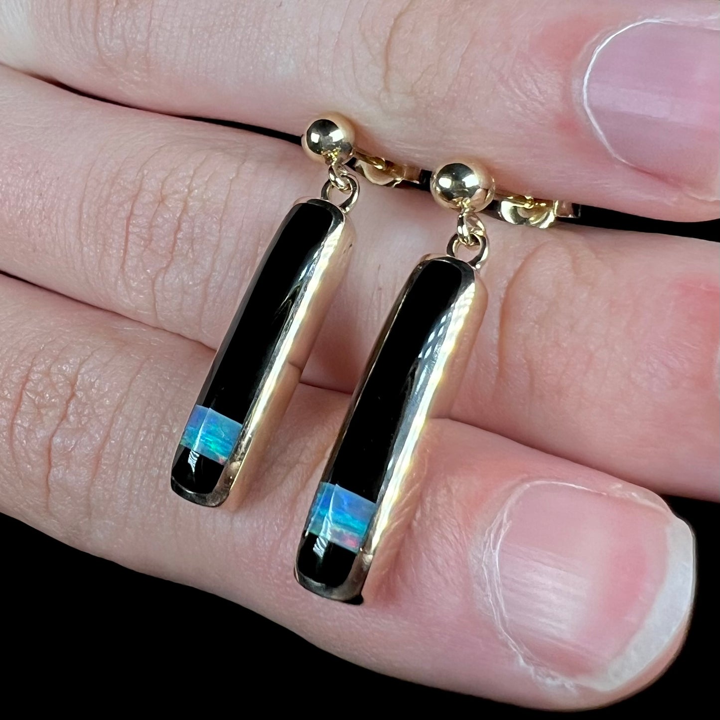 A pair of dangle inlay earrings set with onyx and opal, shown close-up and held between fingers at an angle.