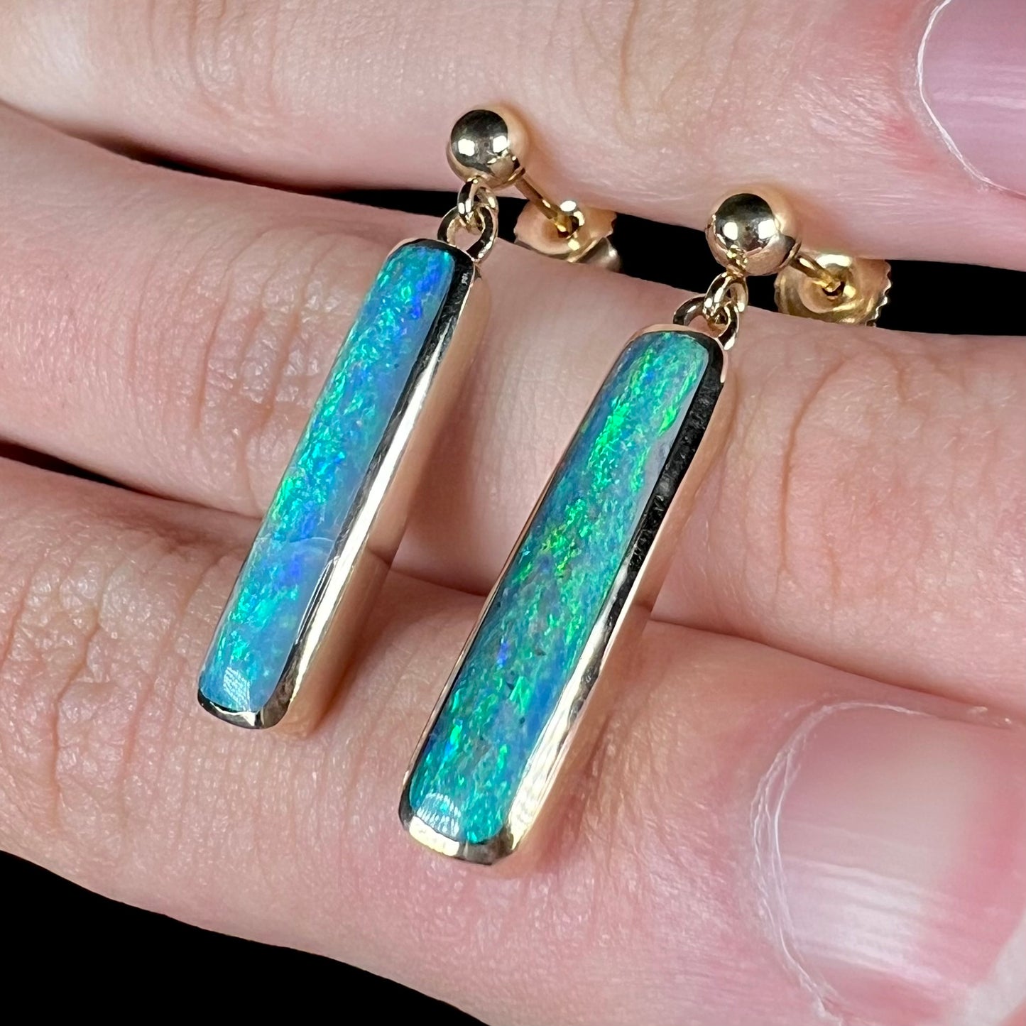 A pair of dark opal dangle earrings with green and blue color play, resting on top of a hand at an angle between fingers.