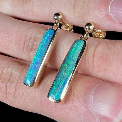 A pair of dark opal dangle earrings with green and blue color play, resting on top of a hand at an angle between fingers.