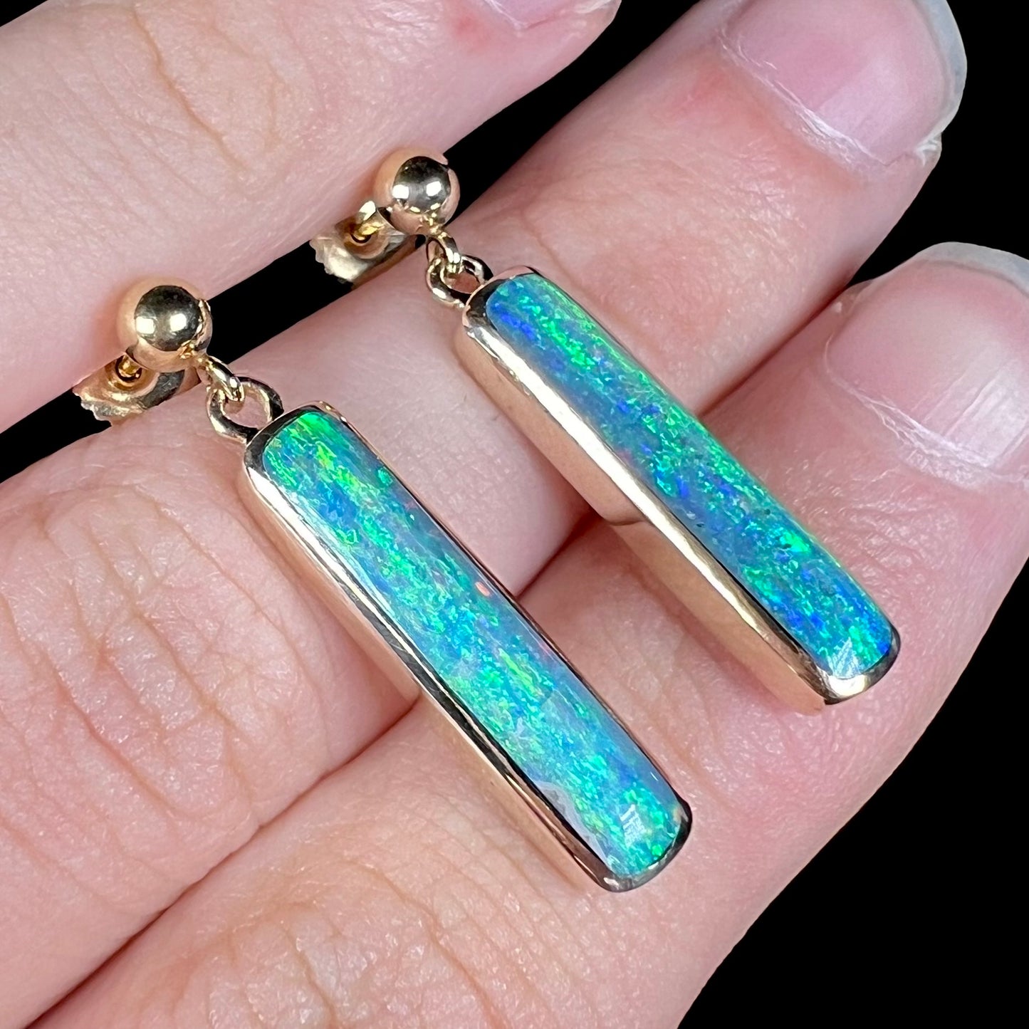 Anlged view of dark opal drop earrings set in yellow gold, shown close-up between two fingers of a hand.