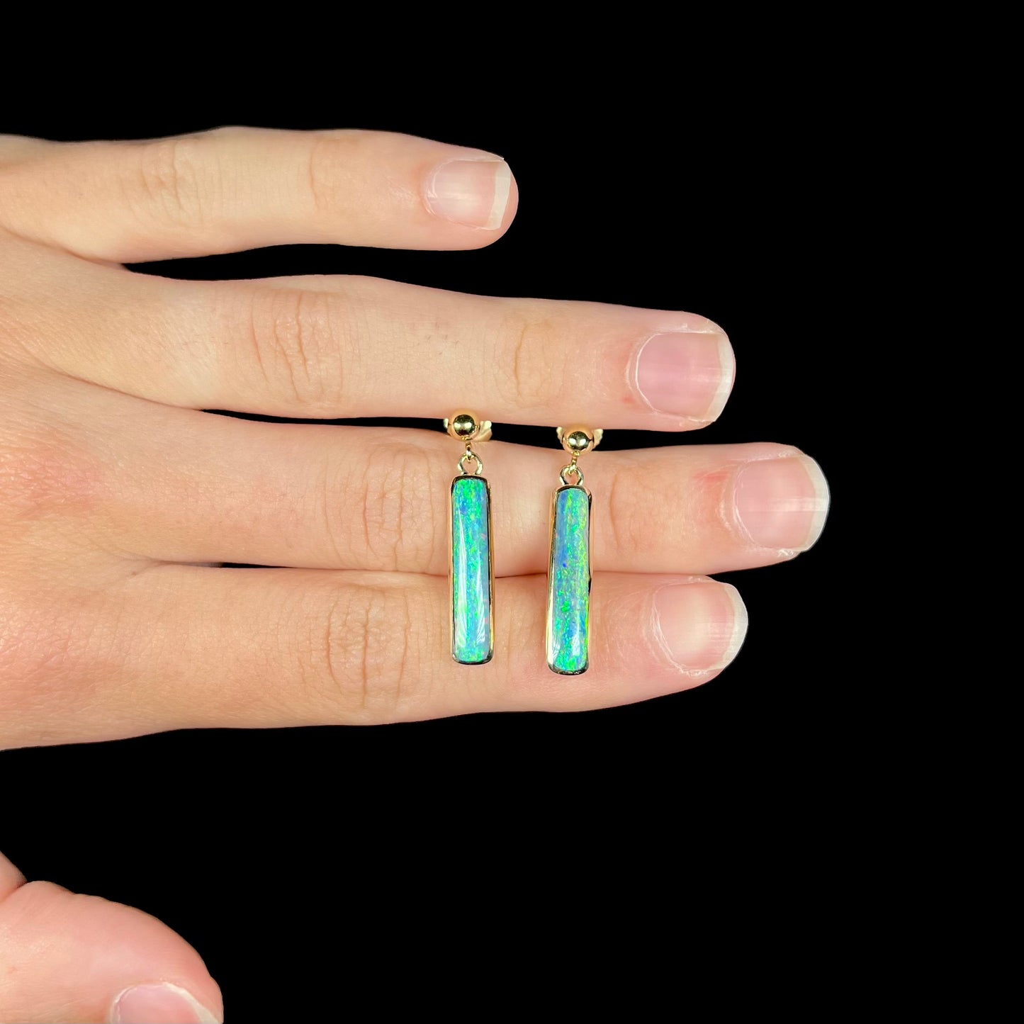A pair of dark opal drop earrings set in yellow gold, held between fingers on top of a hand.