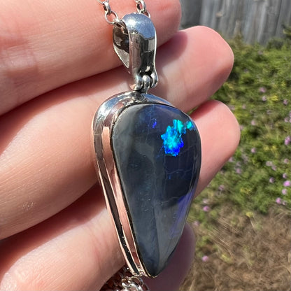 Angled side view of sterling silver Australian dark opal pendant held in sunlight; blue flash of color seen in opal
