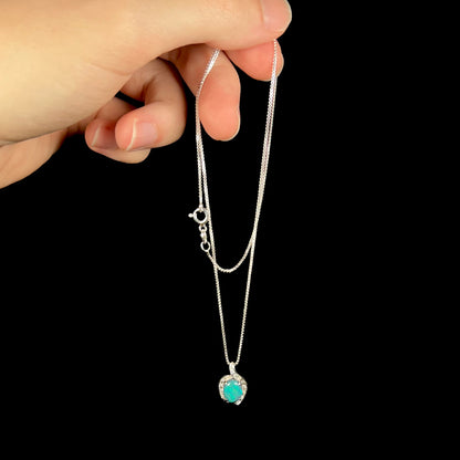 Hand dangling delicate Australian black opal pendant in platinum from silver chain against black background.
