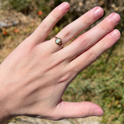 Hand wearing a delicate gold Australian opal and diamond ring in natural light against blurred green and brown background.