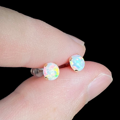 A delicate pair of synthetic opal studs in 14k gold, shown held between two fingers close-up from an angled view.
