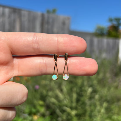 Pair of delicate crystal opal drop earrings in yellow gold held in natural light between two fingers against blurred outdoor backdrop.