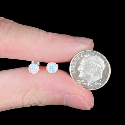 A pair of delicate round 14k gold opal stud earrings, held between two fingers next to a US dime coin for scale.