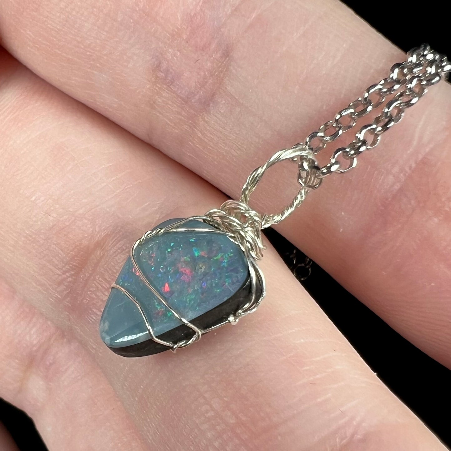 A delicate sterling silver wire wrapped opal doublet women's pendant shown close-up from an angled view, resting on fingers.