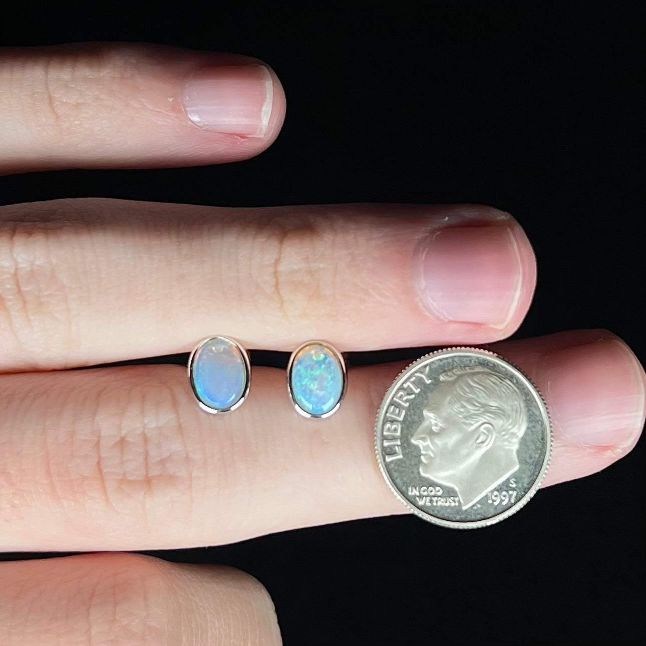 Two delicate-sized white gold Australian opal stud earrings held between two fingers of a hand next to a US dime coin for scale.