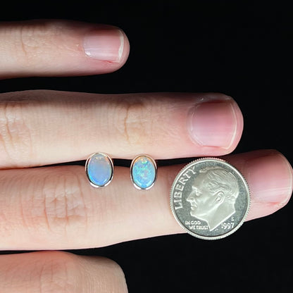 Two delicate-sized white gold Australian opal stud earrings held between two fingers of a hand next to a US dime coin for scale.