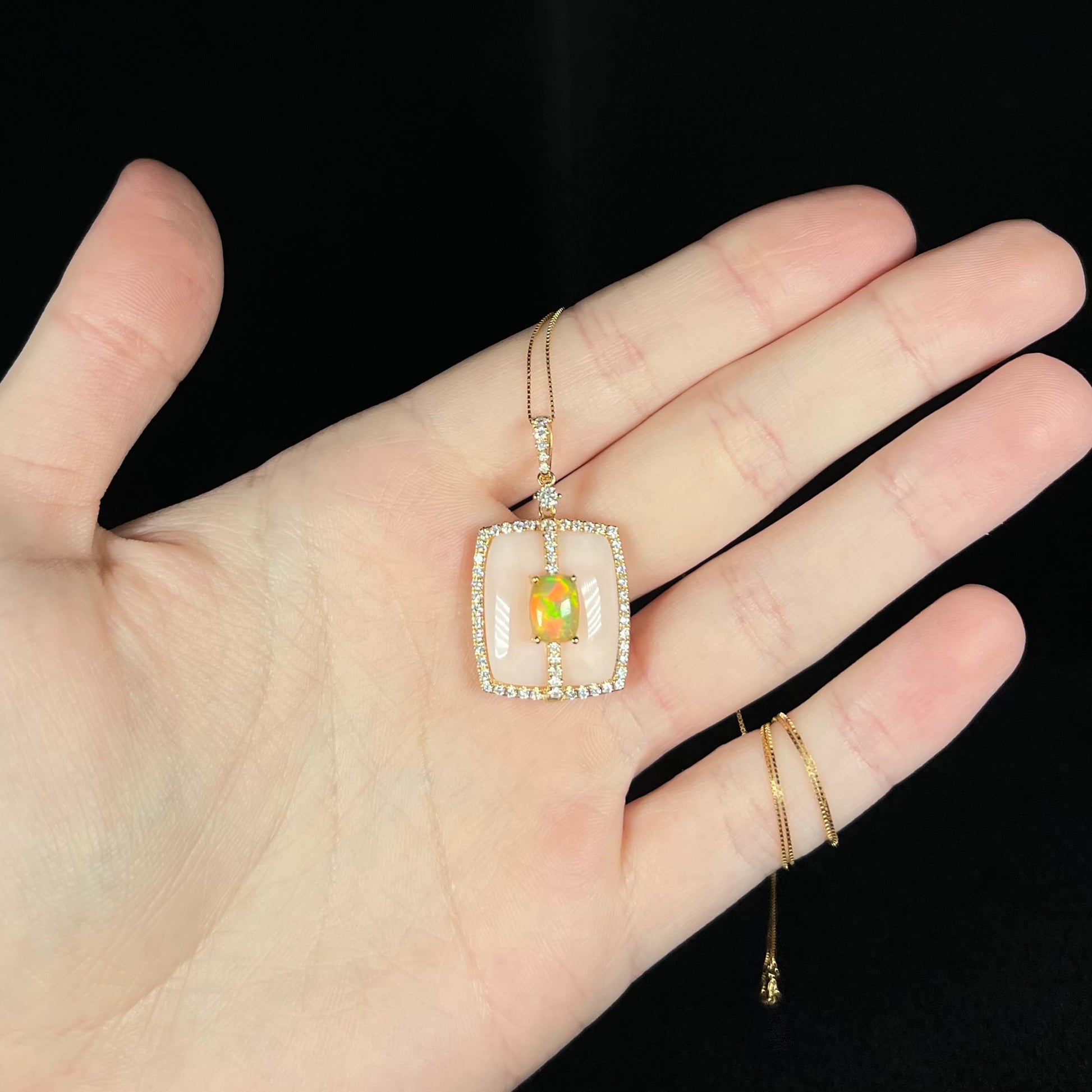 A delicate gold women's fire opal necklace framed with a halo of diamonds, held in a hand against a black background.