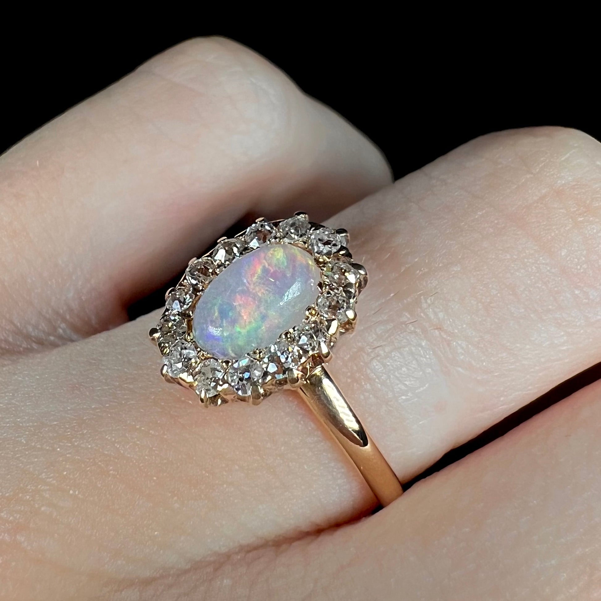 A vintage opal and diamond halo engagement ring on a hand.  The ring is yellow gold, and the opal is pink.