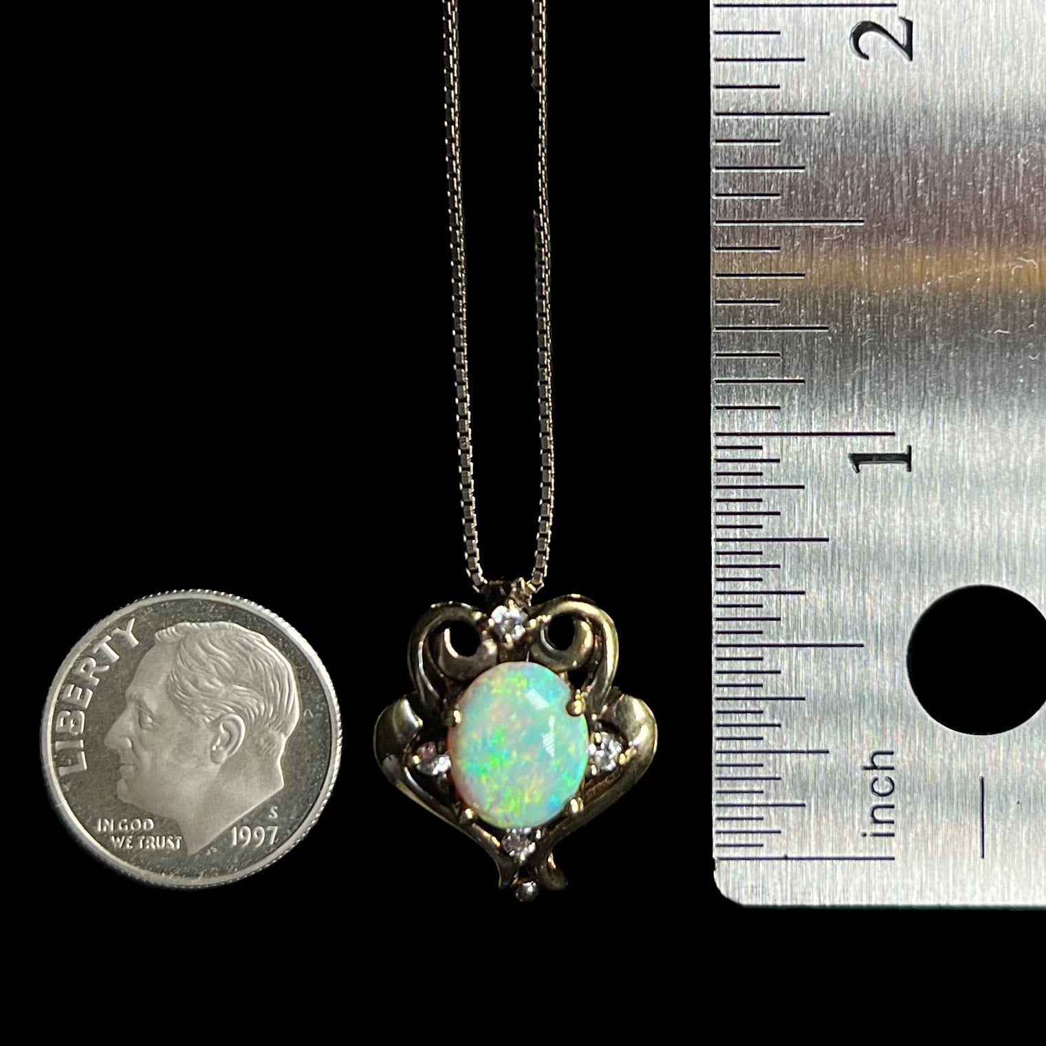 A dime-sized gold-tone Victorian style pendant set with a lab-created opal and CZs, shown on a chain between a US dime coin and ruler for scale.