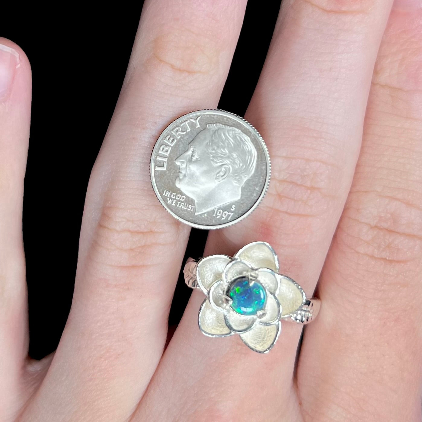 A silver flower-shaped ring with a green iridescent opal center, shown worn on a hand next to a 1997 US dime for scale.