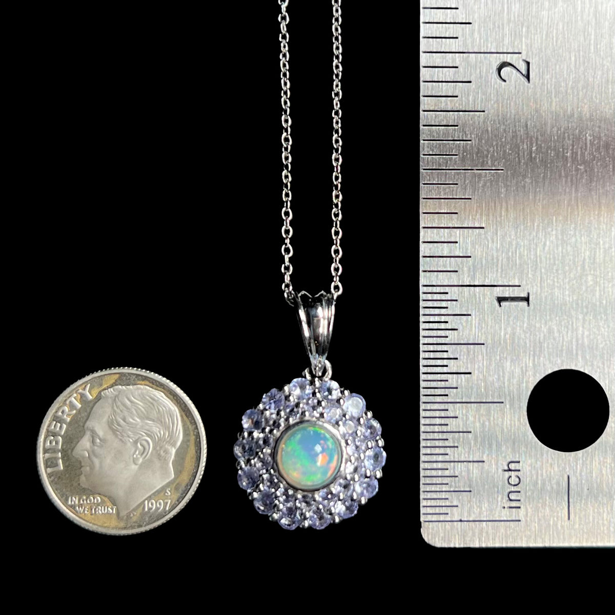 A dime-sized women's sterling silver fire opal and tanzanite halo pendant on a chain between a US dime coin and ruler for scale.