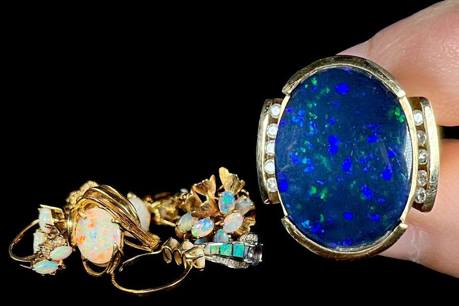 A pile of yellow gold opal jewelry sits behind a large black opal and diamond ring.