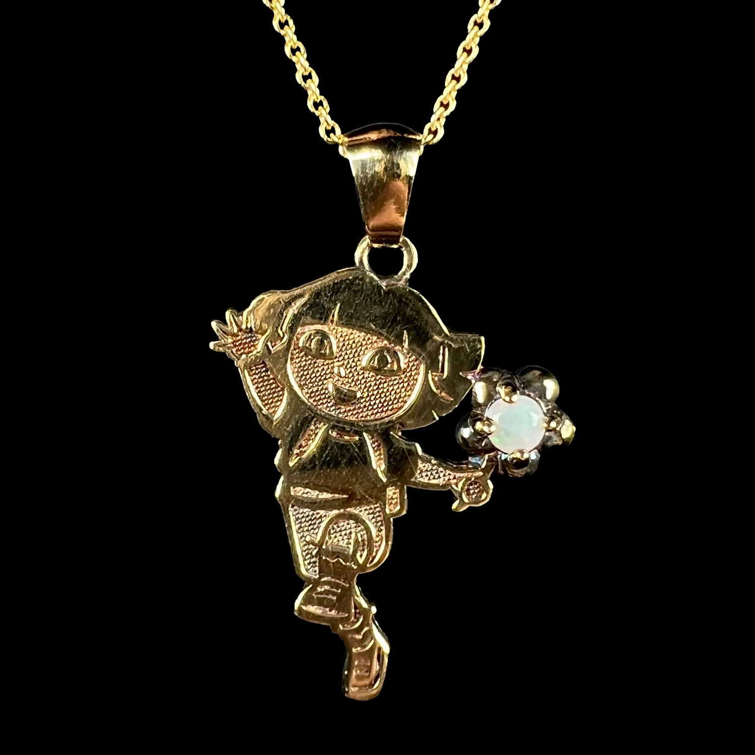 Macro view of gold-plated sterling silver Dora the Explorer necklace with opal flower accent on chain against black background.