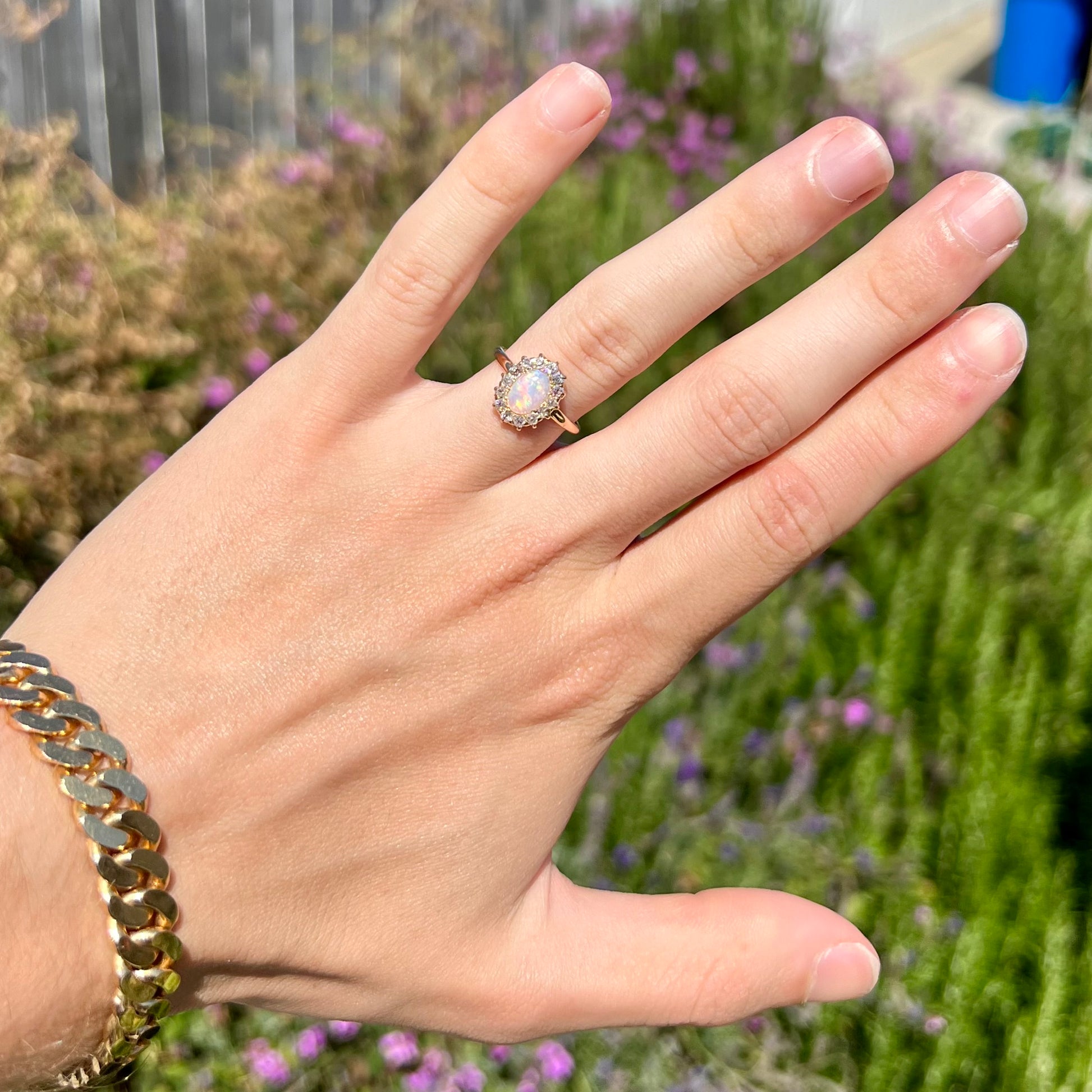 A hand wearing a yellow gold opal and diamond halo engagement ring.  The opal shines pink in the sunlight.