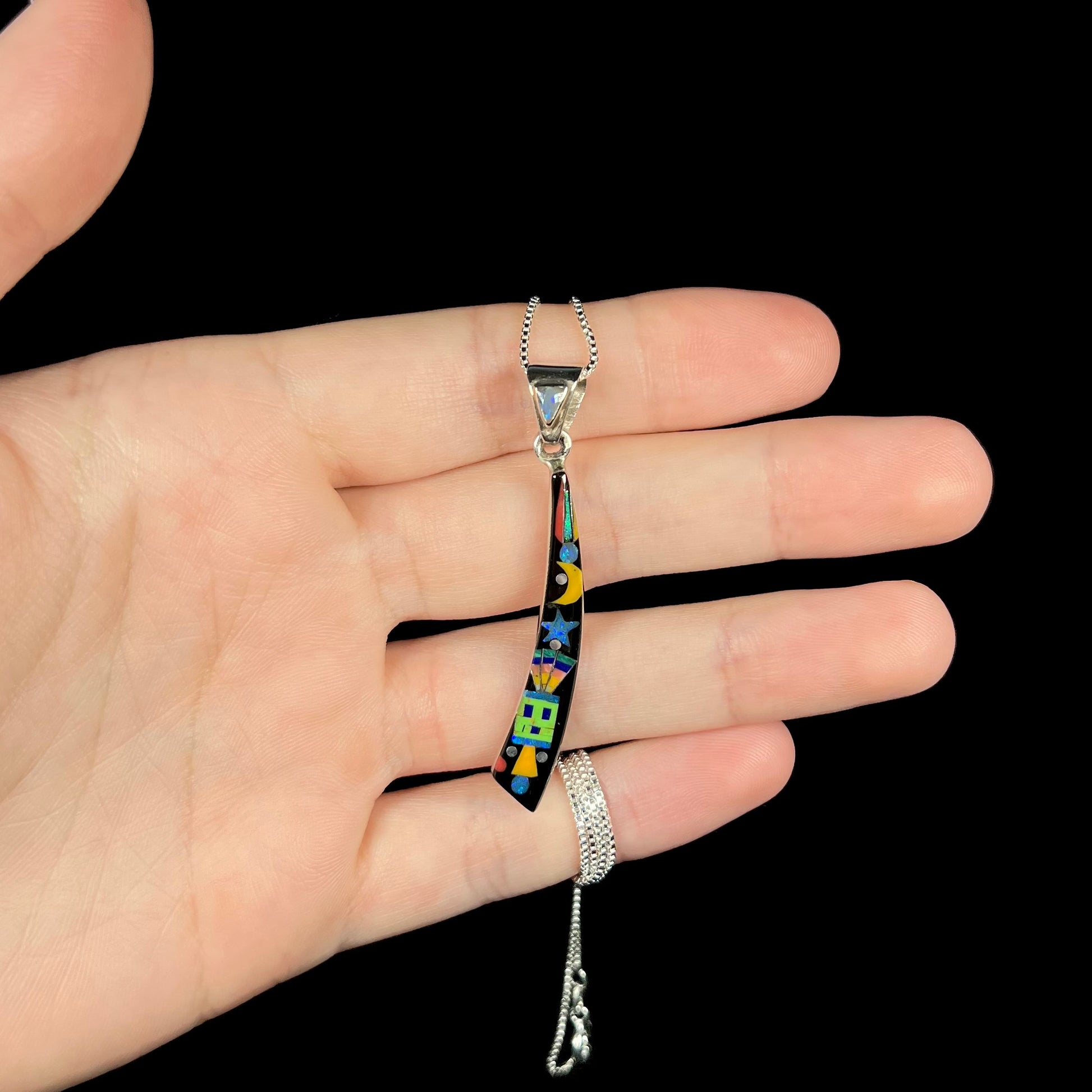 Hand holding men's elongated Southwest-style onyx and gemstone inlay pendant shown on black background.